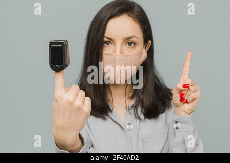 Close up photo female with pulse oximeter on her finger, checks her health. Wears grey dress, isolated grey color background Stock Photo