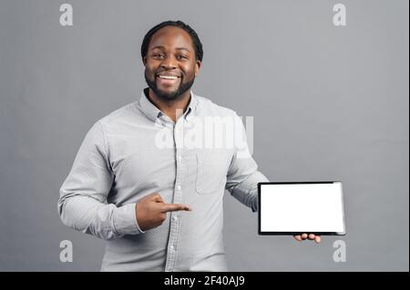 Hilarious African-American young man with dreadlocks holding a digital tablet and point finger at blank screen, template, a black guy presents new app, mockup, isolated on grey background Stock Photo