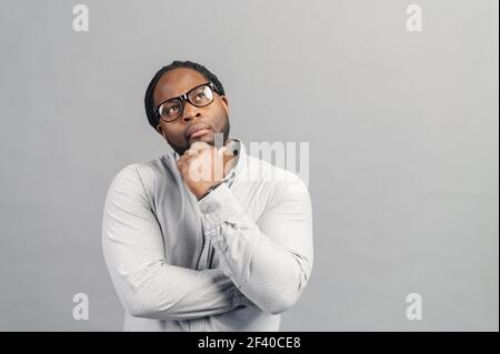 Thoughtful African-American man wearing stylish eyeglasses and smart casual shirt looking new ideas and inspiration, holds chin with a head , looks up, lost in thoughts, isolated on grey Stock Photo