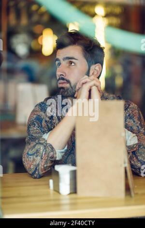 Thoughtful bearded man wearing casual clothes sitting in a modern pub. Guy with beard and modern hairstyle drinking a cola. Stock Photo