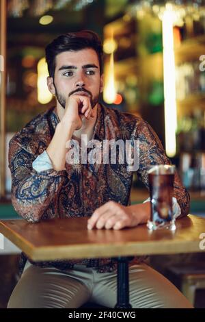 Thoughtful bearded man wearing casual clothes sitting in a modern pub. Guy with beard and modern hairstyle drinking a cola. Stock Photo