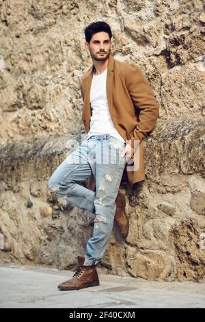 Young man wearing winter clothes in the street. Young bearded guy with modern hairstyle with coat, blue jeans and white t-shirt. Stock Photo