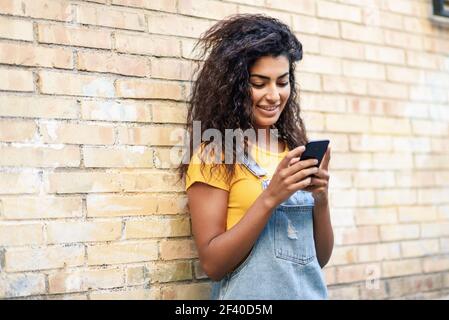 Happy Arab girl using smart phone on brick wall. Smiling woman with curly hairstyle in casual clothes in urban background. Stock Photo