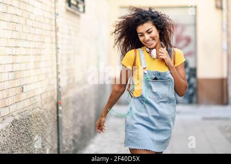 Young African woman with headphones and black curly hairstyle walking outdoors. Happy girl wearing yellow t-shirt and denim dress in urban background. Stock Photo