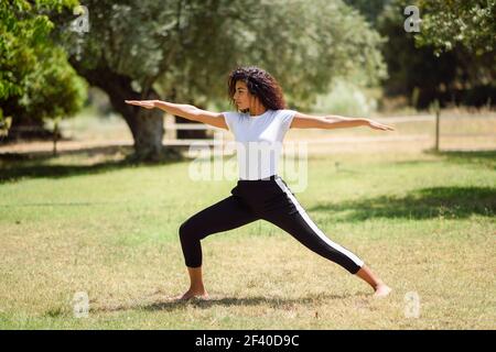 Young Arab woman doing yoga in nature. North African female wearing sport clothes doing Warrior II figure in urban park. Stock Photo