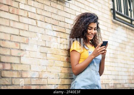 Young North African woman texting with her smart phone outdoors. Smiling Arab girl in casual clothes with black curly hairstyle. Stock Photo