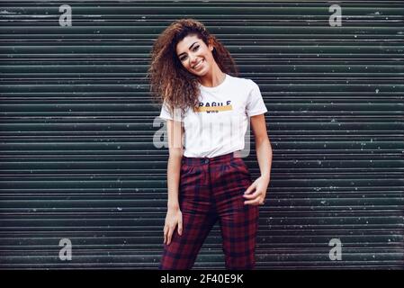 Smiling young arabic woman with black curly hairstyle. Arab girl in casual clothes in the street. Happy female wearing white t-shirt and checked pants against urban blinds. Stock Photo