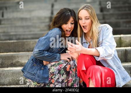 Two young women amazed by what they see on their smart phone outdoors, sitting on urban steps. Friends girls. Stock Photo