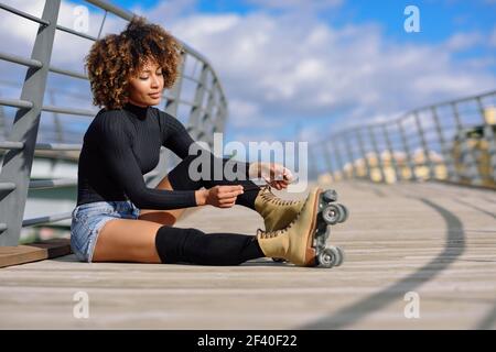 Young smiling black girl sitting on urban bridge and puts on skates. Woman with afro hairstyle rollerblading on sunny day Stock Photo