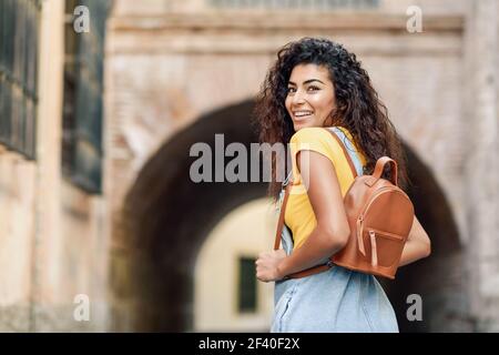 Rear view of young Arab woman with backpack outdoors. Traveler girl in casual clothes in the street. Happy female wearing yellow t-shirt and denim dress in urban background. Stock Photo
