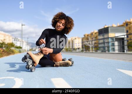 Young smiling black girl sitting on bike line and puts on skates. Woman with afro hairstyle rollerblading on sunny day Stock Photo