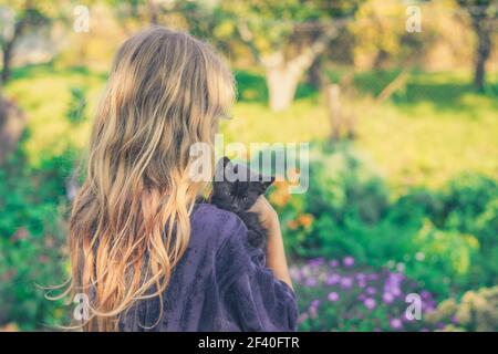 little girl with long blond hair holding black baby cat on her shoulder, back view in the colorful garden during sunny day Stock Photo