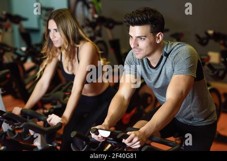 Two people biking in the gym, exercising legs doing cardio workout cycling bikes. Couple in a spinning class wearing sportswear. Stock Photo