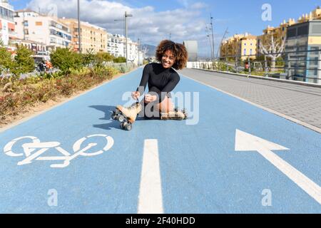 Young smiling black girl sitting on bike line and puts on skates. Woman with afro hairstyle rollerblading on sunny day. Young smiling black girl sitting on bike line and puts on skates. Stock Photo