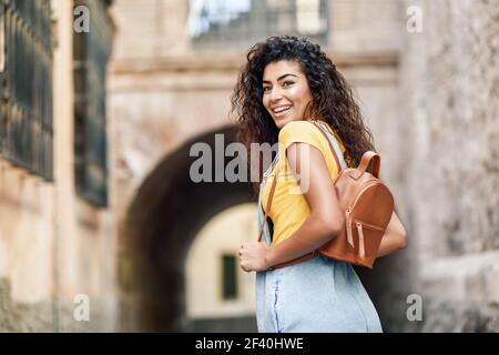 Young North African tourist woman with black curly hairstyle outdoors. Arab traveler girl in casual clothes in the street. Happy female with backpack.. Young African woman with black curly hairstyle outdoors. Stock Photo