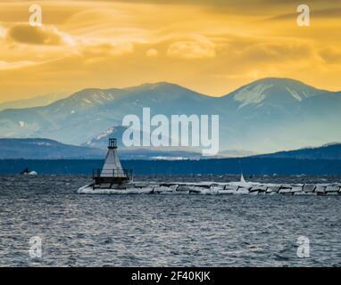 sun setting on Lake Champlain breakwater's lighthouse with the Adirondack mountains in New York across the lake Stock Photo