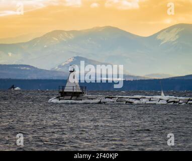 sun setting on Lake Champlain breakwater's lighthouse with the Adirondack mountains in New York across the lake Stock Photo