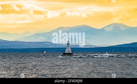 sun setting on Lake Champlain breakwater's lighthouse with the Adirondack mountains in New York across the lake Stock Photo