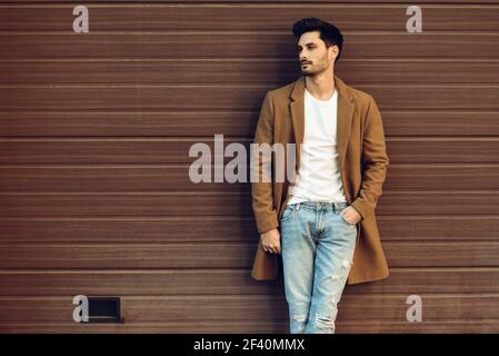 Young man wearing demi-season clothes in the street. Young bearded guy with modern hairstyle with coat, blue jeans and t-shirt against urban wooden blinds.. Young man wearing demi-season clothes in the street. Stock Photo