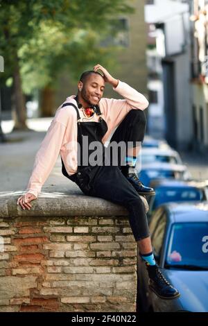 Young black man wearing casual clothes and headphones sitting on urban background. enjoying with eyes closed. Happy African guy with bib pants outdoors. Happy African guy with bib pants outdoors Stock Photo