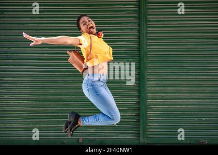 Young African woman jumping on blinds background. Happy girl with very short hair wearing casual clothes.. Young black woman jumping on blinds background. Stock Photo