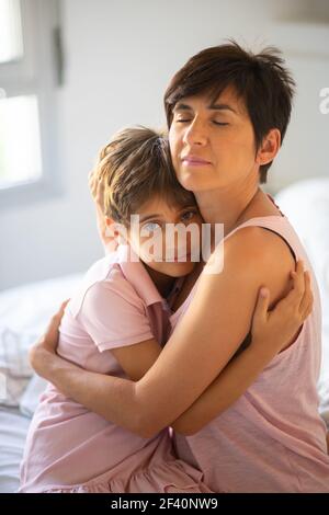 Beautiful young woman and her charming little daughter are hugging on the bed.. Beautiful young woman and her charming little daughter are hugging. Stock Photo