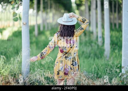 Rear view of hiker young woman, wearing flowered dress, hiking in the countryside.. Young woman, wearing flowered dress, between trees. Stock Photo