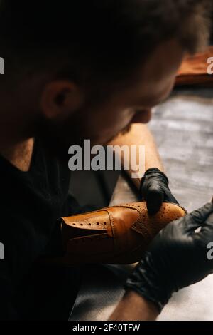 Top view of shoemaker wearing black latex gloves cleaning old light brown leather shoes with rag. Stock Photo