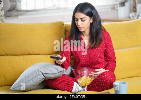 Persian woman at home watching TV and using remote control. Girl eat chips potatoes. Persian woman at home watching TV and using remote control Stock Photo