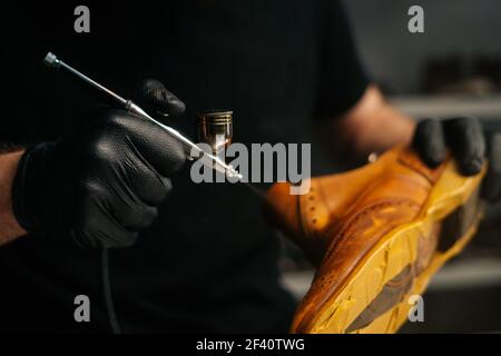 Close-up hands of unrecognizable shoemaker wearing black gloves spraying paint of light brown leather shoes. Stock Photo