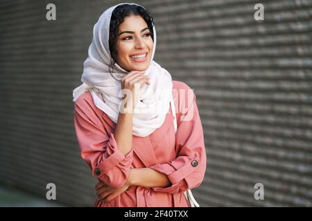 Happy Arab woman wearing hijab headscarf walking in the city center. Muslim female in urban background. Young Muslim woman wearing hijab headscarf walking in the city center. Stock Photo