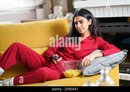 Persian woman at home watching TV and using remote control. Girl eat chips potatoes. Persian woman at home watching TV and using remote control Stock Photo