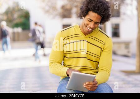 Black man using a digital tablet sitting on a bench outdoors. Guy with afro hair.. Black man using a digital tablet sitting on a bench outdoors. Stock Photo