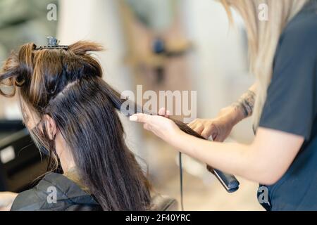 Hairdresser, protected by a mask, making waves in her client&rsquo;s hair with a hair iron in a salon. Business and beauty concepts. Hairdresser, protected by a mask, combing her client&rsquo;s hair with a hair iron in a salon. Stock Photo