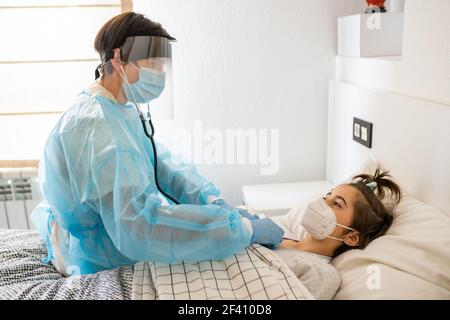 Doctor protected by personal protective equipment examining a little girl with a stethoscope at home.. Doctor examining a little girl with a stethoscope at home. Stock Photo