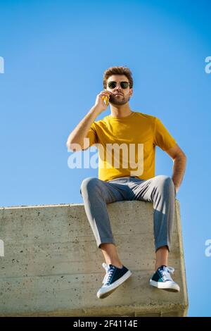 Young man using his smartphone sitting on a ledge outside. Guy wearing sunglasses.. Young man using his smartphone sitting on a ledge outside Stock Photo