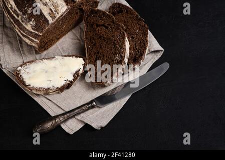 Buttered slice of freshly baked rye bread, table knife on dark background. Artisan sourdough bread. Copy space for recipe. View from above Stock Photo