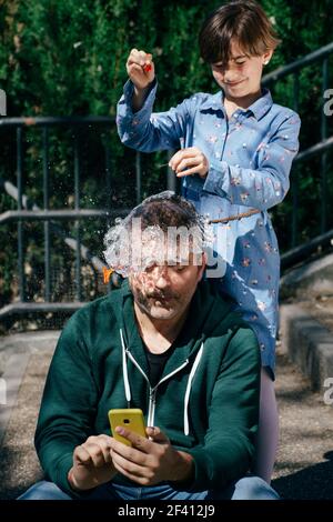 Little girl blowing up a water-filled balloon over her father&rsquo;s head to get him to stop staring at his smartphone.. Girl blowing up a water-filled balloon over her father&rsquo;s head Stock Photo