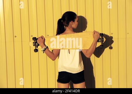 Young girl posing with her skateboard behind her back in front of yellow fence with copyspace, rear view shot. Young girl posing with her skateboard behind her back in front of yellow fence with copyspace, rear view Stock Photo