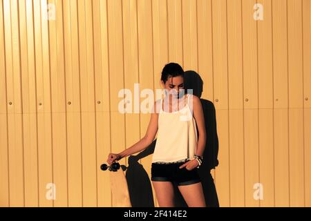 Girl in denim shorts and tank top standing with skateboard in front of yellow mwtal fence and looking down a lot of space for text. Girl in denim shorts and tank top standing with skateboard in front of yellow mwtal fence and looking down Stock Photo