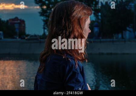 Red haired girl looking at night river in city park with highlight on her pretty ginger hair. Lonely weekend.. Red haired girl looking at night river in city park with highlight on her pretty ginger hair. Stock Photo