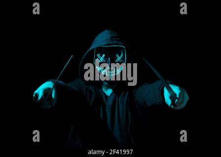 Man armed with two machetes and wearing a scary lighting neon glow mask and a hoodie on black background. Stock Photo
