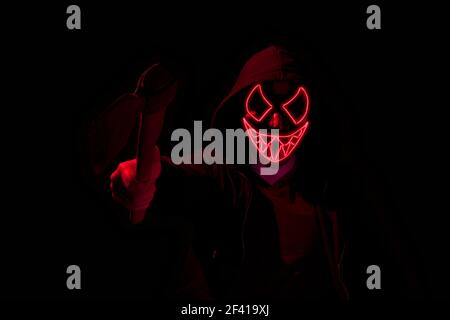 Man armed with an axe and wearing a scary lighting neon glow mask and a hoodie on black background. Stock Photo