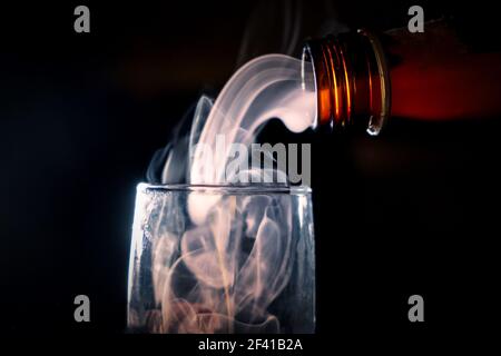 The neck of a brown bottle with alcohol overlooked a glass, from it comes an evil spirit that fills the glass. Photo on a black background with a copyspace.. The neck of a brown bottle with alcohol overlooked a glass, from it comes an evil spirit that fills the glass. Stock Photo
