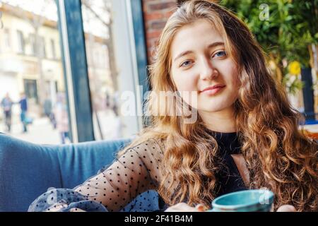 Red-haired girl with blue eyes sits in a cafe and looks into the camera over the table with cups. Red-haired girl with blue eyes sits in a cafe and looks into the camera Stock Photo
