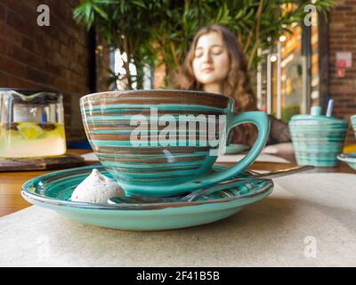 A red-haired girl sits at a table in a cafe blurry image shot, in the foreground a ceramic cup of green color with a pattern. A red-haired girl sits at a table in a cafe blurry image, in the foreground of a ceramic cup Stock Photo