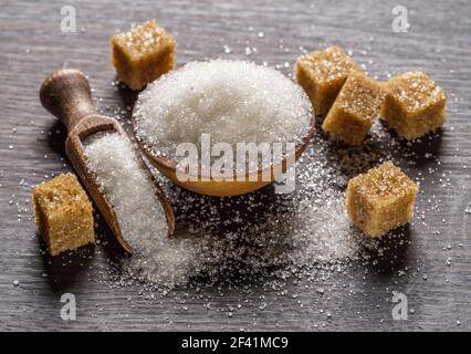 Granulated table sugar in wooden bowl and in the spoon and sugar cubes around it on the table. Stock Photo