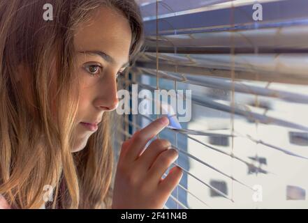 Young girl looking through window blinds, looking sad Stock Photo