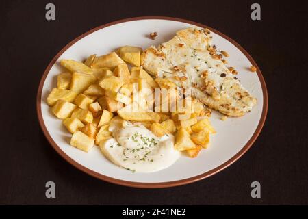 Dish with a hake fillet with garlic and parsley accompanied by aioli sauce and potatoes on a dark wooden table. Food and plating. Stock Photo