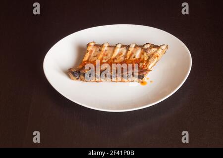 Isolated white plate with some juicy pork ribs cooked in a spicy vegetable sauce on a dark wooden table. Homemade food with tasty meat. Stock Photo